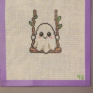 May include: A needlepoint canvas featuring a white ghost with black eyes and pink cheeks sitting on a brown wooden swing suspended by two brown ropes with small green leaves. The canvas has a purple border and the letters GS in the bottom corner.