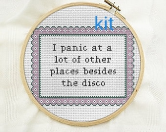 May include: A circular wooden embroidery hoop containing white fabric with a cross-stitch design. The text reads, I panic at a lot of other places besides the disco, surrounded by a decorative border in shades of purple, green, and black.
