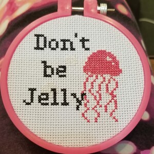 May include: A pink and white embroidery hoop with a cross-stitch design of a pink jellyfish and the text "Don't be Jelly".