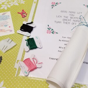 May include: A cross-stitch kit with a white Aida cloth, floss in black, green, and pink, and needles. The kit includes instructions for a cross-stitch design with the text "Good Moms Let You Lick The Beaters Great Moms Turn Them Off".