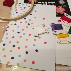 Puede incluir: Un kit de punto de cruz con fondo blanco y lunares de colores. El kit incluye un aro de bordado de madera, hilo de bordar blanco y cinco carretes de hilo de bordar DMC en rojo, morado, amarillo, verde y blanco.
