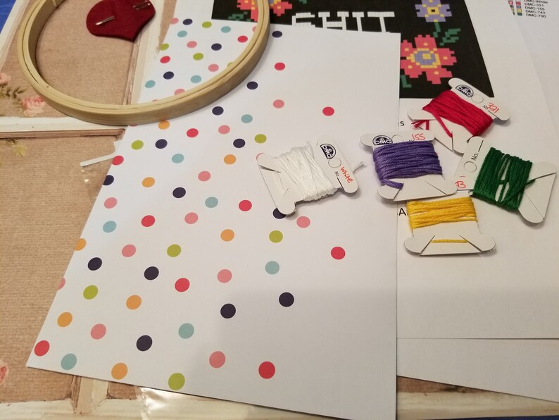 Puede incluir: Un kit de punto de cruz con fondo blanco y lunares de colores. El kit incluye un aro de bordado de madera, hilo de bordar blanco y cinco carretes de hilo de bordar DMC en rojo, morado, amarillo, verde y blanco.