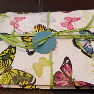 K&ouml;nnte beinhalten: Eine wei&szlig;e Geschenkbox, verpackt in Papier mit Schmetterlingsmuster, mit einem gr&uuml;nen Band und einem blauen runden Anh&auml;nger. Die Box ist mit einem gr&uuml;nen Band gebunden und hat einen blauen runden Anh&auml;nger.