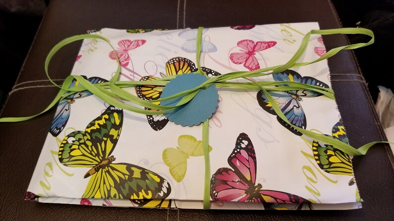 K&ouml;nnte beinhalten: Eine wei&szlig;e Geschenkbox, verpackt in Papier mit Schmetterlingsmuster, mit einem gr&uuml;nen Band und einem blauen runden Anh&auml;nger. Die Box ist mit einem gr&uuml;nen Band gebunden und hat einen blauen runden Anh&auml;nger.