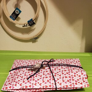 May include: A white gift bag with a red heart pattern is tied with a black ribbon. The bag is sitting on a green surface.