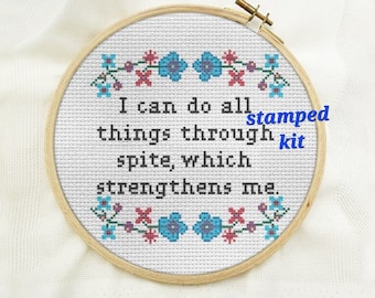 May include: Cross-stitch embroidery in a wooden hoop. The white fabric features a quote in black text: "I can do all things through spite, which strengthens me." Floral border in blue, red, and purple. The words "stamped kit" are in blue.