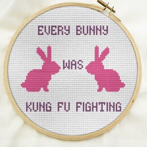 May include: A white embroidery hoop with a pink cross-stitch design. The design features two pink bunnies facing each other with the text "EVERY BUNNY WAS KUNG FU FIGHTING".