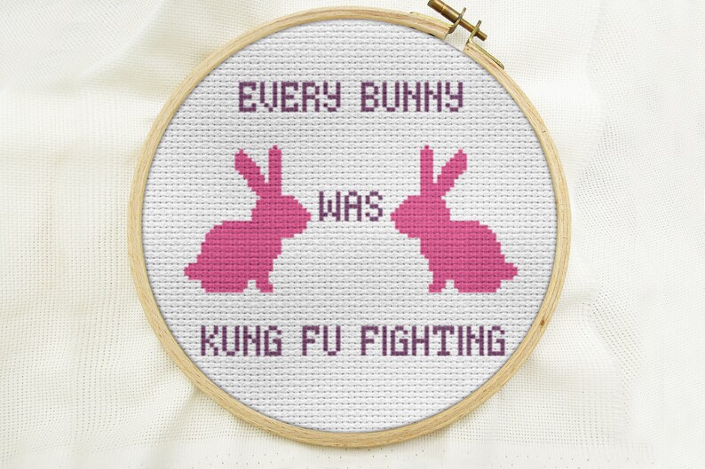 May include: A white embroidery hoop with a pink cross-stitch design. The design features two pink bunnies facing each other with the text "EVERY BUNNY WAS KUNG FU FIGHTING".