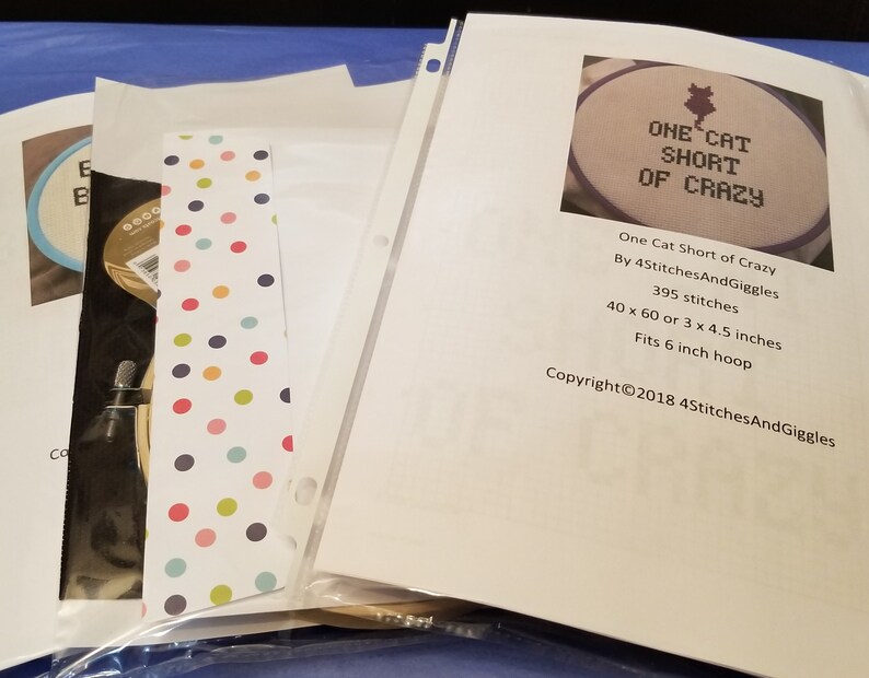 Puede incluir: Una colecci&oacute;n de patrones de punto de cruz, incluido uno titulado "One Cat Short of Crazy". Los patrones est&aacute;n en fundas de pl&aacute;stico transparente, con un marcap&aacute;ginas de lunares de colores. El texto indica que se adapta a un bastidor de 15 cm y tiene 395 puntadas.