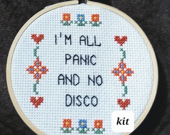May include: A circular wooden embroidery hoop containing white Aida fabric with cross-stitched text that reads I'M ALL PANIC AND NO DISCO. The design features red and blue flowers, green stems, and red hearts. A small white label says kit.