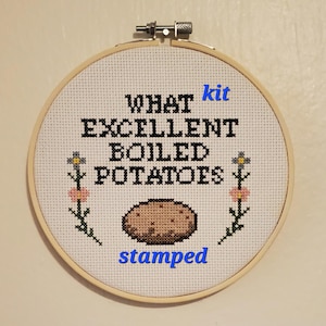 May include: Cross-stitch embroidery in a wooden hoop. The design features the text "WHAT kit EXCELLENT BOILED POTATOES stamped" in black and blue. A brown potato is in the center, with floral accents on each side.