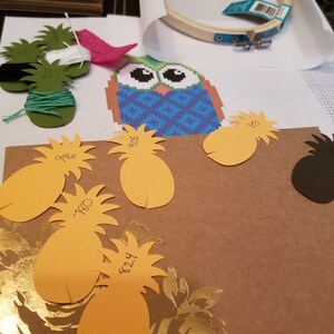 K&ouml;nnte beinhalten: Nahaufnahme eines Bastelprojekts mit einem blauen und gr&uuml;nen Eulen-Kreuzstichmuster, einem h&ouml;lzernen Stickrahmen und mehreren orangenen Papier-Ananas mit darauf geschriebenen Zahlen.