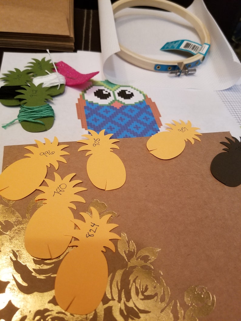 K&ouml;nnte beinhalten: Nahaufnahme eines Bastelprojekts mit einem blauen und gr&uuml;nen Eulen-Kreuzstichmuster, einem h&ouml;lzernen Stickrahmen und mehreren orangenen Papier-Ananas mit darauf geschriebenen Zahlen.