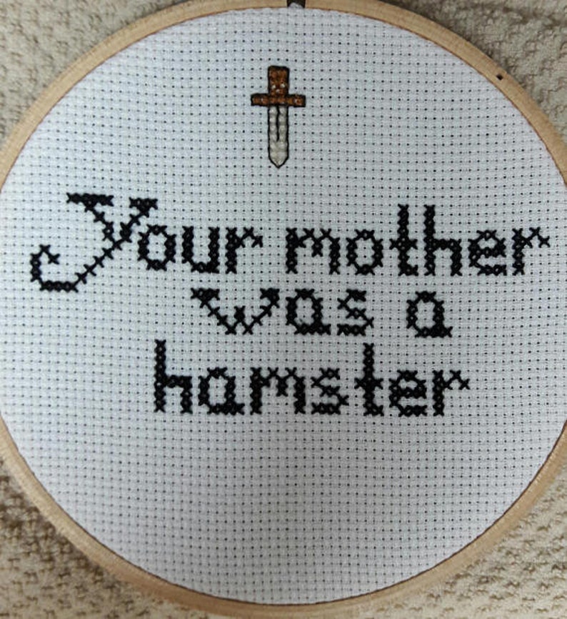 Puede incluir: Bastidor de bordado de punto de cruz con la frase "Your mother was a hamster" en hilo negro. Una peque&ntilde;a espada bordada est&aacute; encima del texto. La tela es blanca y el bastidor es de madera.