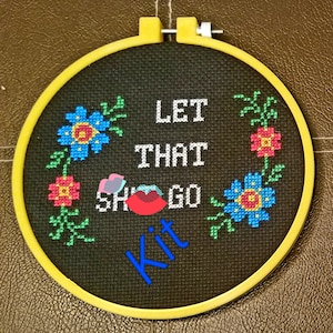 May include: A black embroidery hoop with a yellow frame. The embroidery says "Let that sh*t go" with a red lip and flowers in the corners. The word "Kit" is written in blue below the text.
