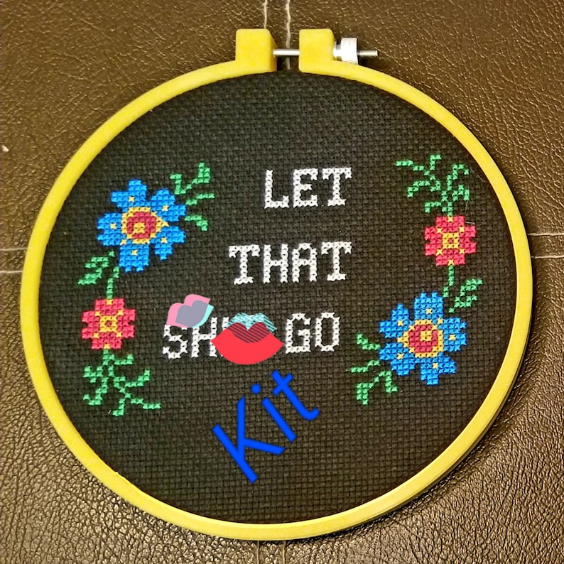 May include: A black embroidery hoop with a yellow frame. The embroidery says "Let that sh*t go" with a red lip and flowers in the corners. The word "Kit" is written in blue below the text.
