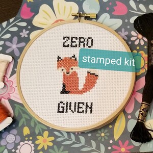 May include: A cross-stitch embroidery kit featuring a fox with the text "Zero Given" on a white fabric in a wooden embroidery hoop. The kit is labeled as a "stamped kit".