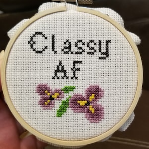 May include: A white embroidery hoop with a cross-stitch design that says "Classy AF" in black thread. The design also includes two purple flowers with yellow centers.