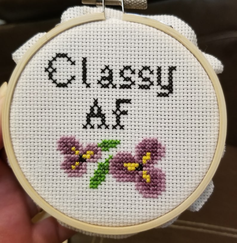 May include: A white embroidery hoop with a cross-stitch design that says "Classy AF" in black thread. The design also includes two purple flowers with yellow centers.