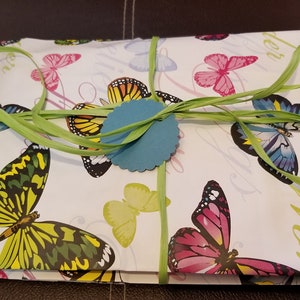 Puede incluir: Una caja de regalo blanca envuelta en papel con estampado de mariposas con una cinta verde y una etiqueta redonda azul. La caja est&aacute; atada con una cinta verde y tiene una etiqueta redonda azul.
