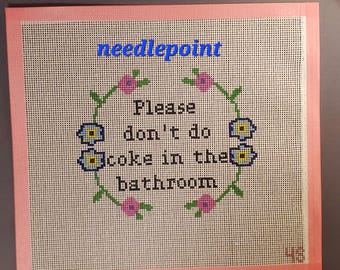 May include: A needlepoint canvas featuring a floral wreath design in pink, green, and blue surrounding the black text "Please don't do coke in the bathroom". The canvas has a pink border and the word "needlepoint" in blue at the top.