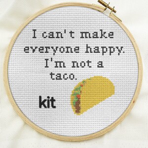 May include: White embroidery hoop with a cross-stitch design that reads "I can't make everyone happy. I'm not a taco." and a yellow taco with green and red fillings. The words "kit" are printed below the design.
