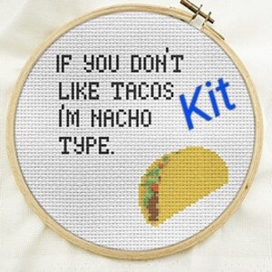 May include: A white embroidery hoop with a cross-stitch design that says "If you don't like tacos I'm nacho type."  The design also includes a yellow taco with green and red details. The word "Kit" is written in blue letters.