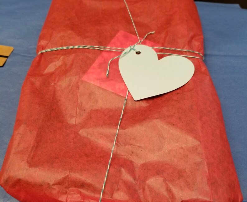 Peut inclure: Un sac cadeau rouge ficel&eacute; avec de la ficelle et une &eacute;tiquette blanche en forme de c&oelig;ur. Le sac est en papier de soie et a une texture froiss&eacute;e.