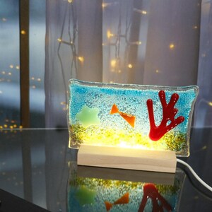 May include: A decorative glass lamp featuring an underwater scene with a blue background, orange fish, a red coral, and a green fish. The lamp sits on a wooden base and emits a warm yellow light. The lamp is connected to a white power cord.