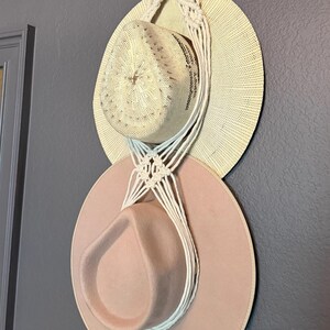 May include: A macrame wall hanger made of off-white cord featuring a circular wooden ring at the top, designed to hold two hats. The top hat is a light beige woven straw style, and the bottom hat is a solid, smooth, light pink felted hat.