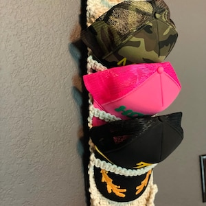 May include: A macrame hat organizer with four baseball caps. The caps are in various colors, including camouflage, hot pink, black, and black with gold embroidery. The organizer is cream-colored and hangs against a gray wall.