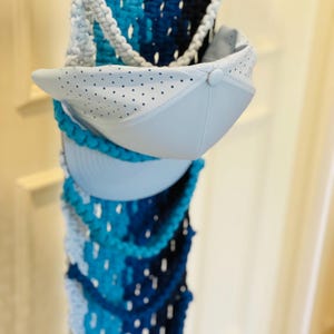 May include: A pale blue baseball cap with a perforated design is shown. The cap is in front of a woven column with shades of blue and white. The column has a decorative, rope-like trim. The woven column is made of a textured material.
