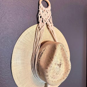 May include: A beige straw hat suspended by a cream-coloured macrame cord hanger. The hanger features a circular wooden ring at the top, with intricate knotted patterns extending downwards to cradle the hat, ending in a short fringe at the bottom.