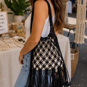 May include: A black macrame tote bag featuring an intricate knotted net pattern, long shoulder straps, and a series of decorative tassels along the bottom edge. The bag contains a solid off-white fabric pouch insert to hold items securely inside.