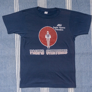 May include: A navy blue t-shirt with a red circle and a graphic of Uncle Sam. The text "ASU Lyric Opera Theatre" and "PACIFIC OVERTURES" are printed on the shirt.