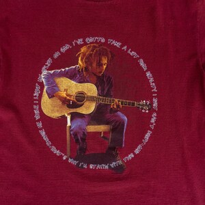 May include: A red t-shirt with a circular graphic featuring a man playing an acoustic guitar. The text around the graphic reads "I've gotta take a lift from reality, just can't stay with this body long. Why I'm stayin' with that's me while I light my life on God."