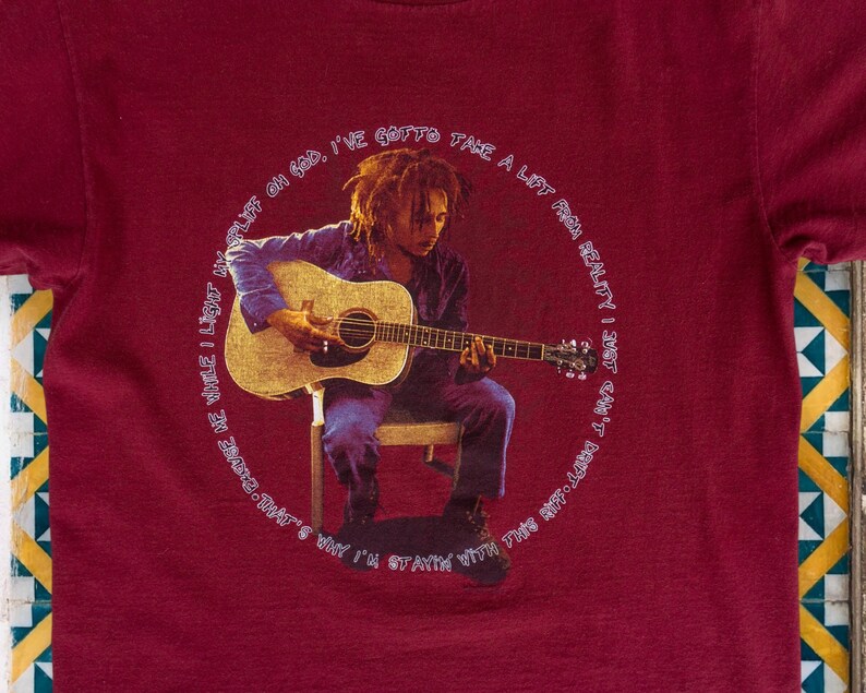 May include: A red t-shirt with a circular graphic featuring a man playing an acoustic guitar. The text around the graphic reads "I've gotta take a lift from reality, just can't stay with this body long. Why I'm stayin' with that's me while I light my life on God."