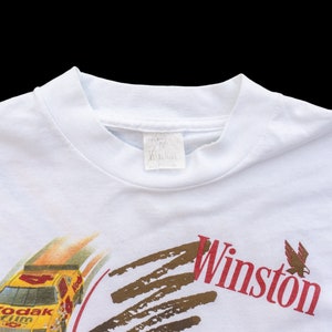 May include: White t-shirt with a yellow race car and the words "Kodak film" on the front. The car is driving on a brown track with the word "Winston" in red letters above it.