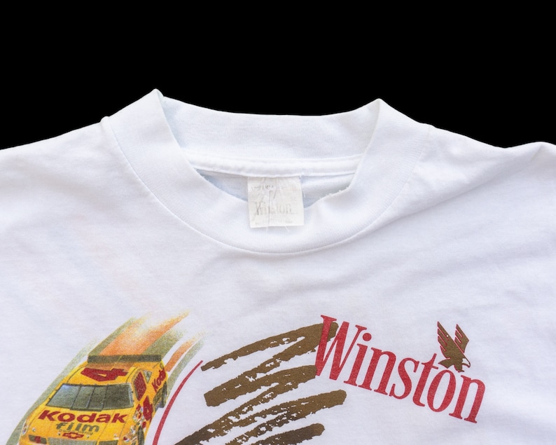 May include: White t-shirt with a yellow race car and the words "Kodak film" on the front. The car is driving on a brown track with the word "Winston" in red letters above it.