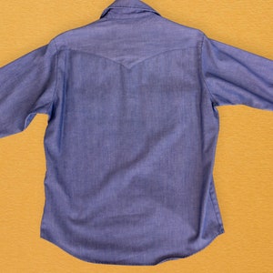 May include: A blue denim shirt with a pointed collar and long sleeves. The shirt has a single button closure at the top and a yoke at the back.
