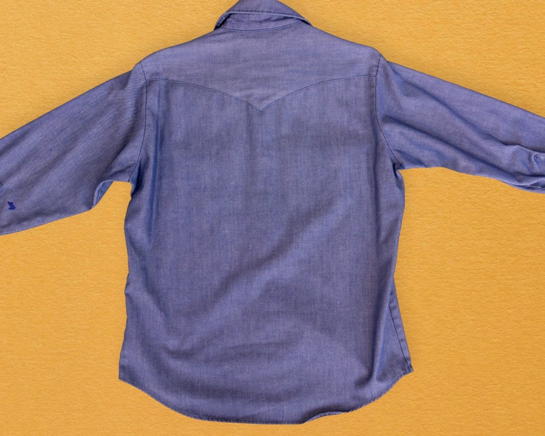 May include: A blue denim shirt with a pointed collar and long sleeves. The shirt has a single button closure at the top and a yoke at the back.