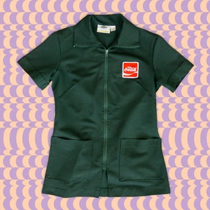 May include: A dark green uniform dress with a zipper closure and two pockets. The dress has short sleeves and a small red Coca-Cola logo patch on the front.
