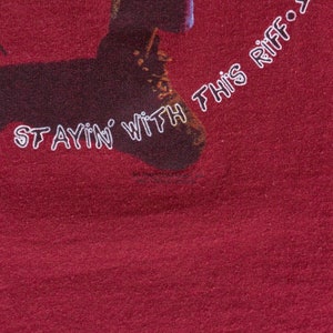 May include: A maroon t-shirt with a graphic of a person's legs wearing brown boots. The text "I'm stayin' with this riff..." is printed on the shirt.