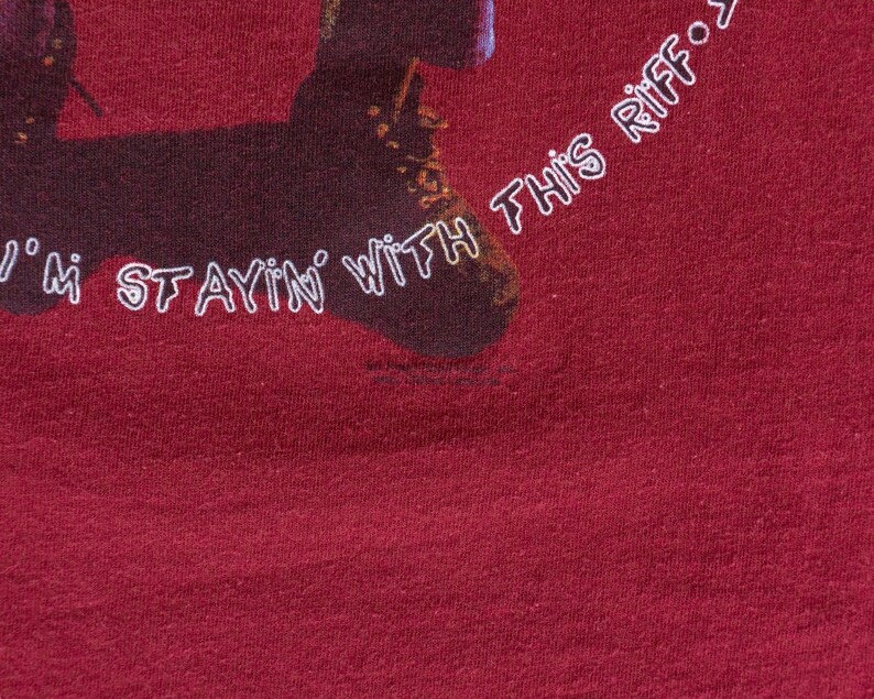 May include: A maroon t-shirt with a graphic of a person's legs wearing brown boots. The text "I'm stayin' with this riff..." is printed on the shirt.