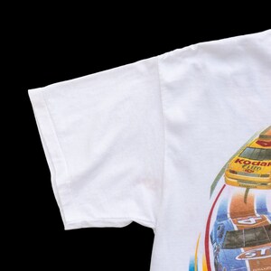 May include: White t-shirt with a graphic print of two race cars on a track. The cars are yellow and blue with the words "Kodak Film" and "STP Grand Prix" on them.