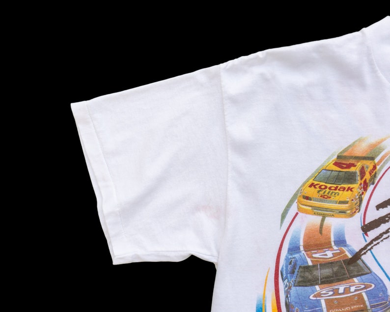 May include: White t-shirt with a graphic print of two race cars on a track. The cars are yellow and blue with the words "Kodak Film" and "STP Grand Prix" on them.