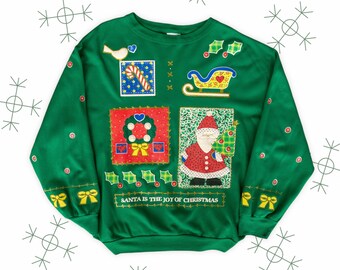 Op de afbeelding: Groene kersttrui met een patchwork-ontwerp met een vogel, een snoepstok, een krans, de kerstman en een slee. De trui heeft de tekst "SANTA IS THE JOY OF CHRISTMAS" onderaan.