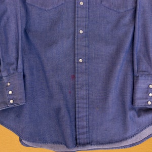 May include: A blue denim long-sleeve shirt with a button-down front and a single chest pocket. The shirt has a faded appearance and a few small stains.