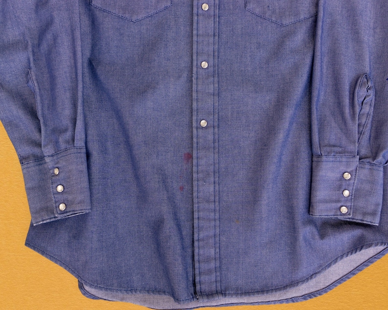 May include: A blue denim long-sleeve shirt with a button-down front and a single chest pocket. The shirt has a faded appearance and a few small stains.