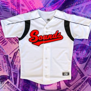 May include: White baseball jersey with black trim and a red "Sounds" logo on the front.